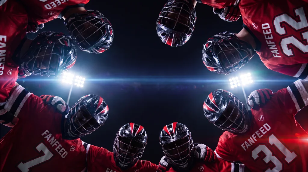 Looking up from inside a lacrosse team huddle at night, the players' helmets ringing the frame against the stadium floodlights.